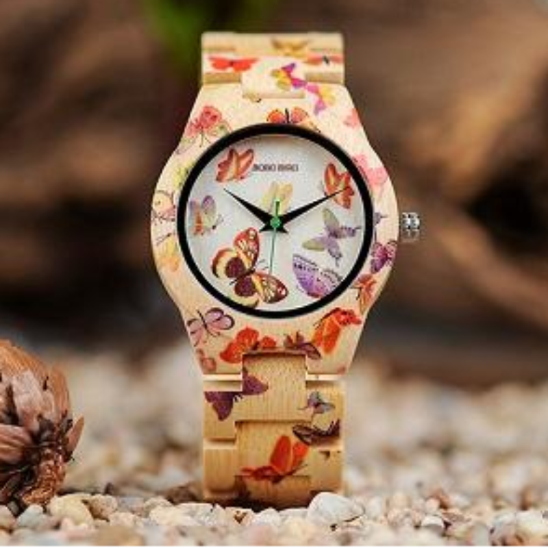 A wristwatch with a light wood-grain strap and case decorated with colorful butterfly and floral patterns, displayed on a bed of small pebbles with a blurred natural background.