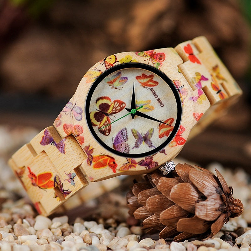 Wooden watch with butterfly design on strap, placed on a pine cone with a natural background.