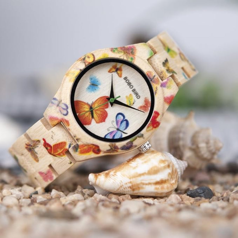 A wooden watch with a colorful butterfly print on the case and strap, resting on seashells over small pebbles. The watch face is white with black hands and the brand name 'BOBO BIRD' is visible.