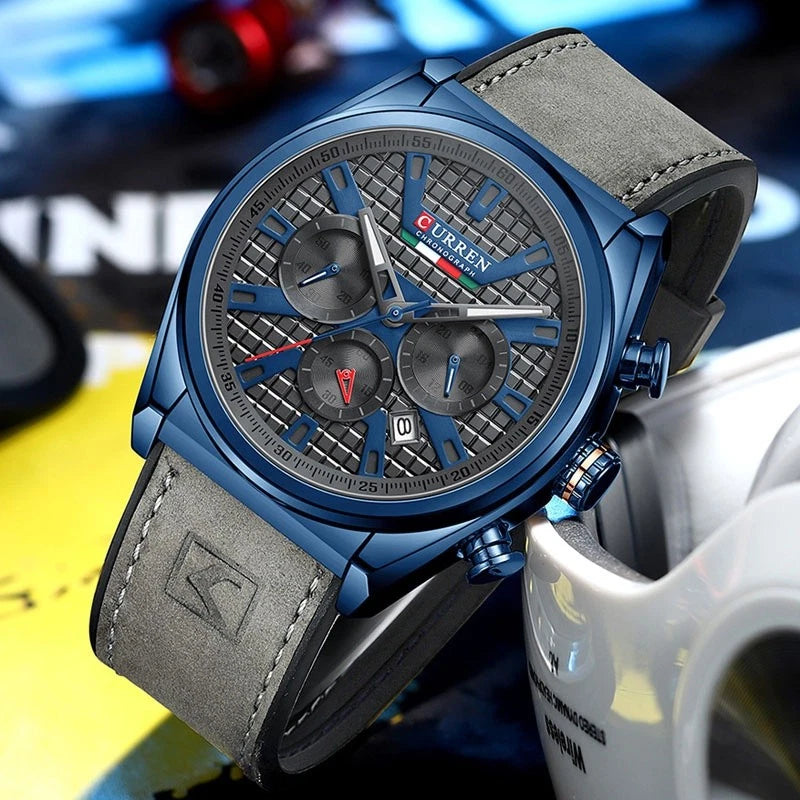 A men's wristwatch with a blue metal case, textured dark gray dial, and gray leather strap, photographed close-up against a blurred multicolor background.
