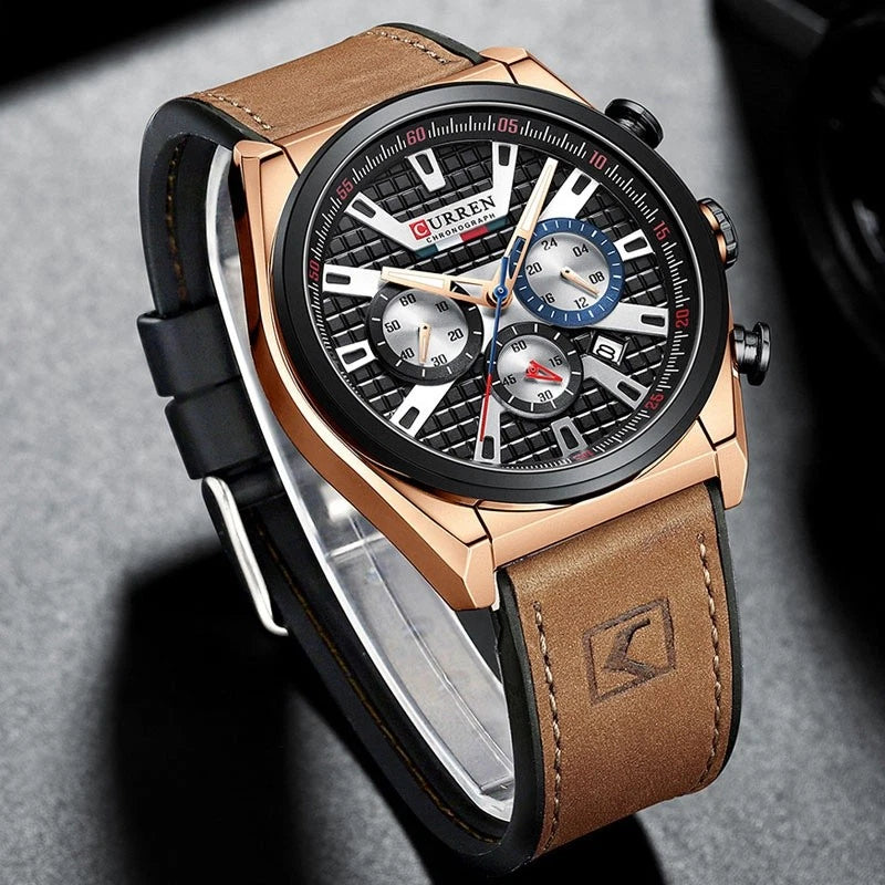 A luxury chronograph wristwatch with a black textured dial, rose gold-toned case, and a brown leather strap, displayed on a gray textured surface.