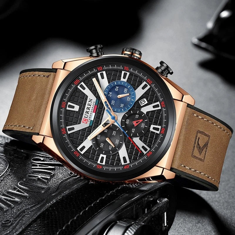 A luxury men's chronograph watch with a rose gold-tone case, black textured dial, brown leather strap, and blue sub-dials, resting on a black leather surface.