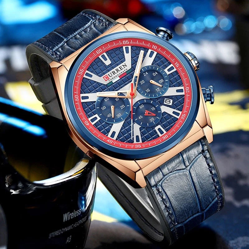 A close-up of a wristwatch with a rose gold-tone case, blue textured dial, red outer ring, and dark blue leather strap, photographed against a blurred blue and yellow background.