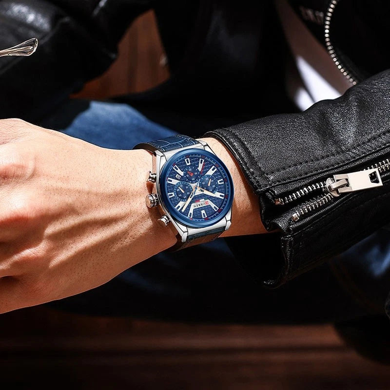 A men's wristwatch with a blue dial and blue strap, worn on a person's wrist. The watch has a silver-tone case and silver-tone indices, and the background shows a person wearing a black leather jacket.