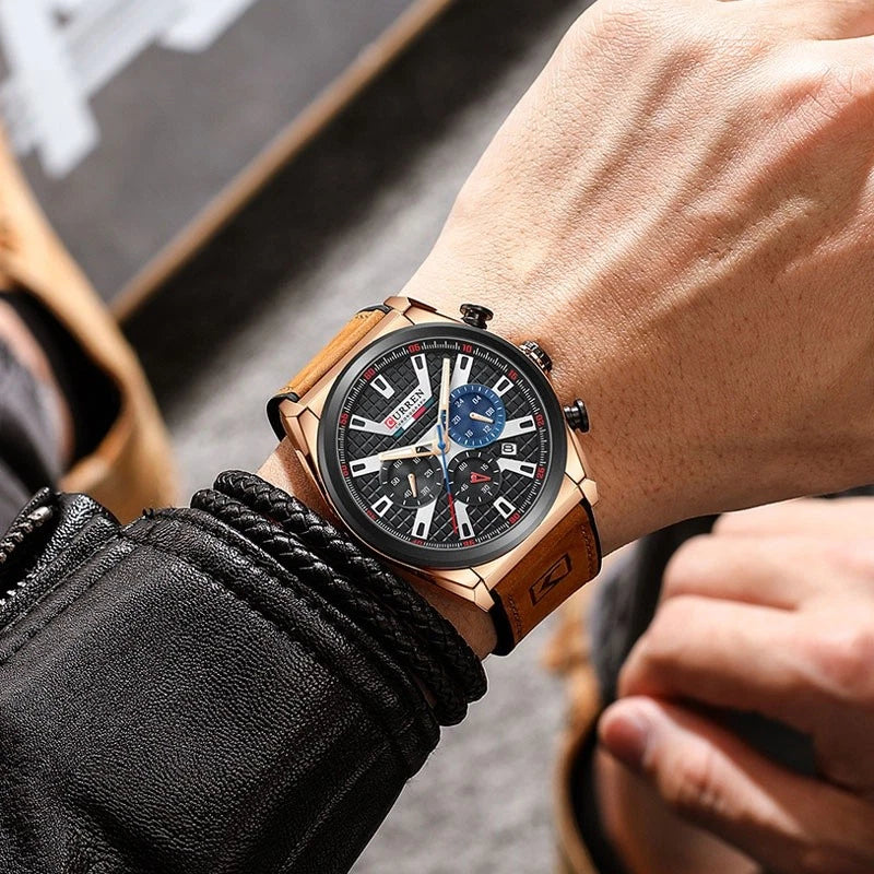A men's sport watch with a black dial, rose gold-toned case, and brown leather strap, shown worn on a wrist.