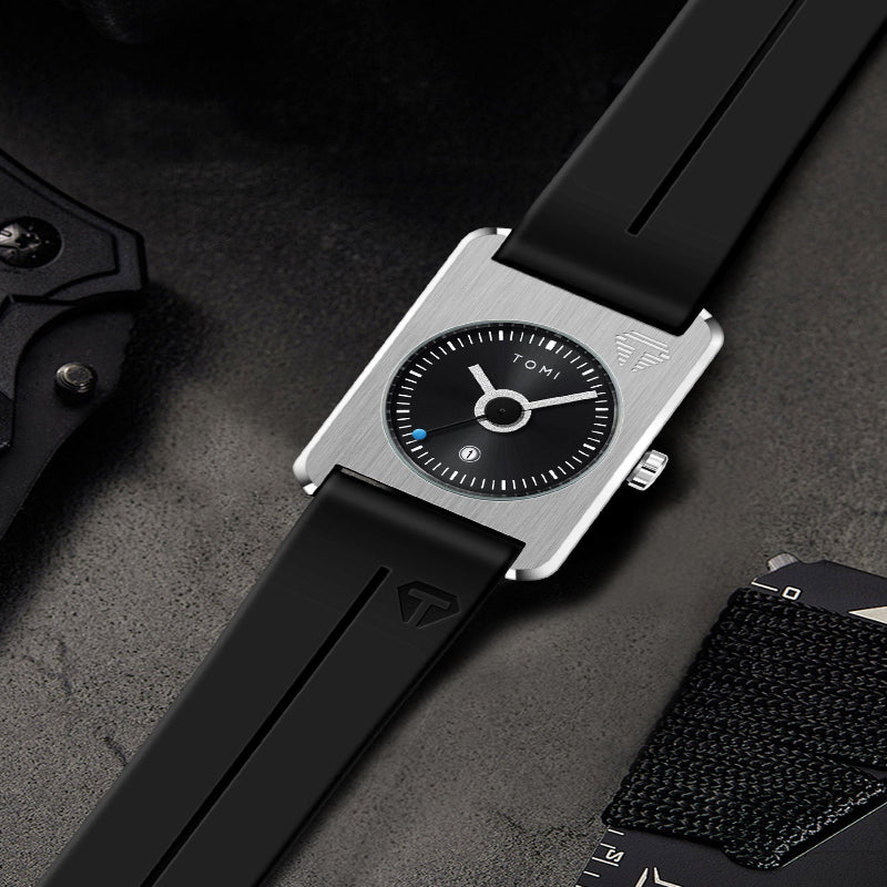 A Tomi analog watch with a silver-toned rectangular case, black dial, and black strap, styled diagonally on a dark textured surface with a knife and card nearby.