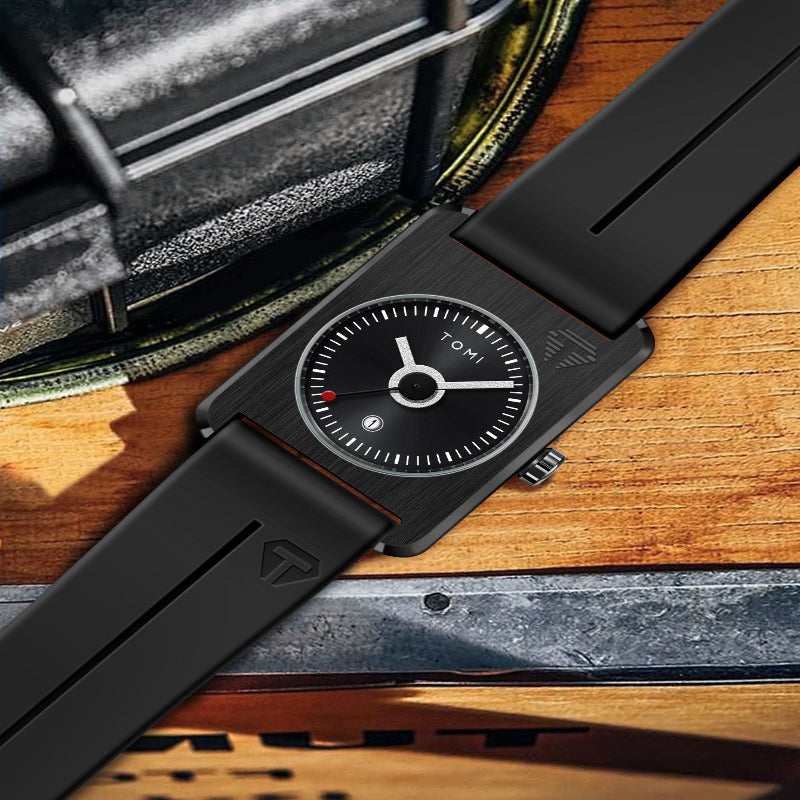 A black rectangular Tomi watch with a matte black strap, shown diagonally on a wooden surface next to a dark textured case.