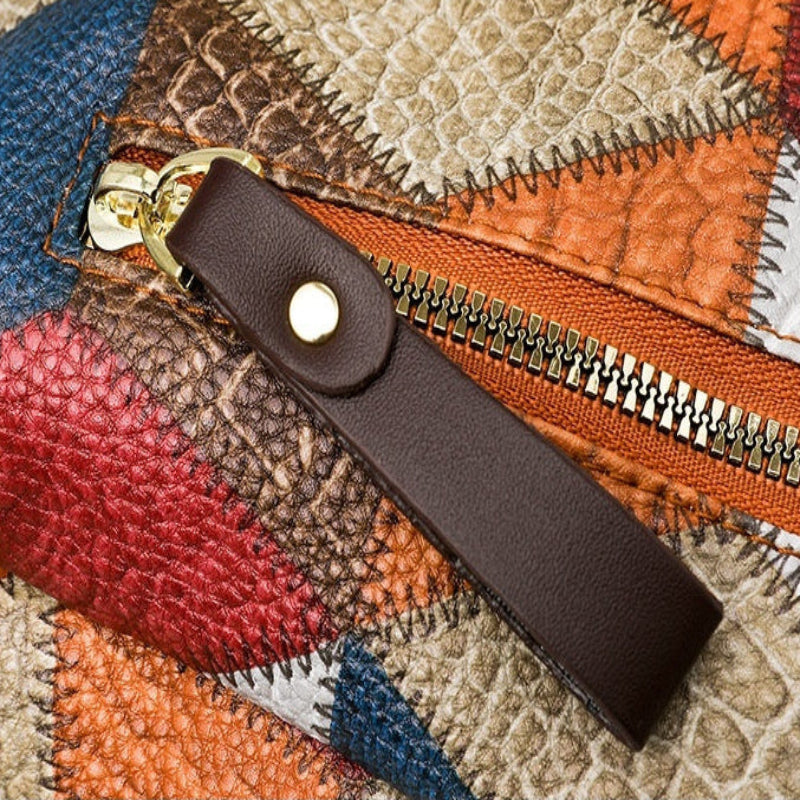 A close-up of a color-block leather zipper pull attached to a multicolor patchwork-textured bag. The zipper pull is dark brown leather with a gold-tone snap button and ring.
