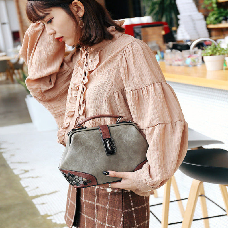 A woman in a cafe setting wearing a beige long-sleeve blouse and holding a gray top-handle handbag with brown leather trim and a decorative beaded strap detail.