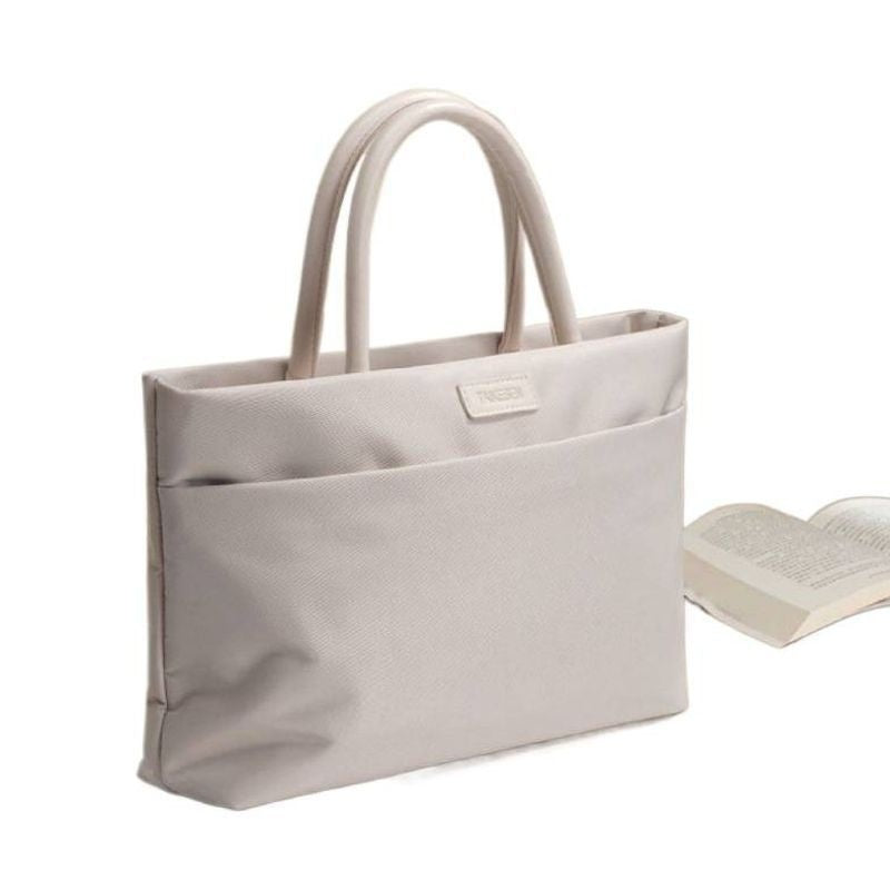 A light beige canvas tote bag with dual top handles and a front slip pocket, shown next to an open book on a white background.