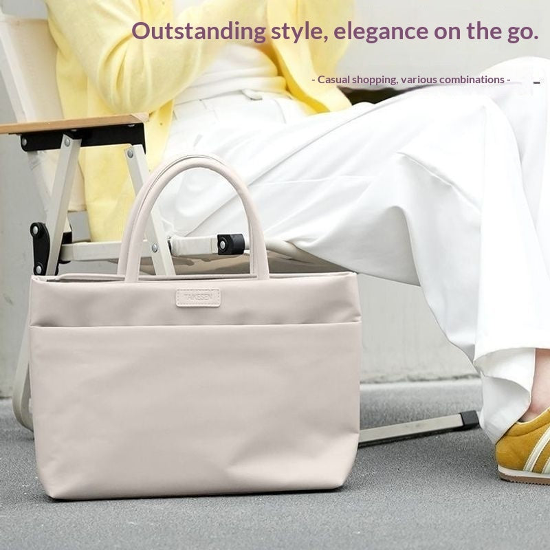 A light beige tote bag with dual top handles sits on a gray floor next to a person sitting on a folding chair. The person is wearing a yellow cardigan, white pants, and yellow shoes.