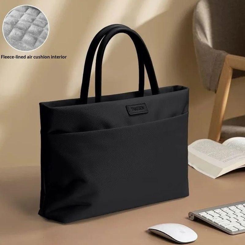 A black tote bag with dual top handles and a front pocket, shown on a desk with a mouse, keyboard, and open book. A circular inset in the upper left shows the quilted cushioned interior lining, with text reading 'Fleece-lined air cushion interior.